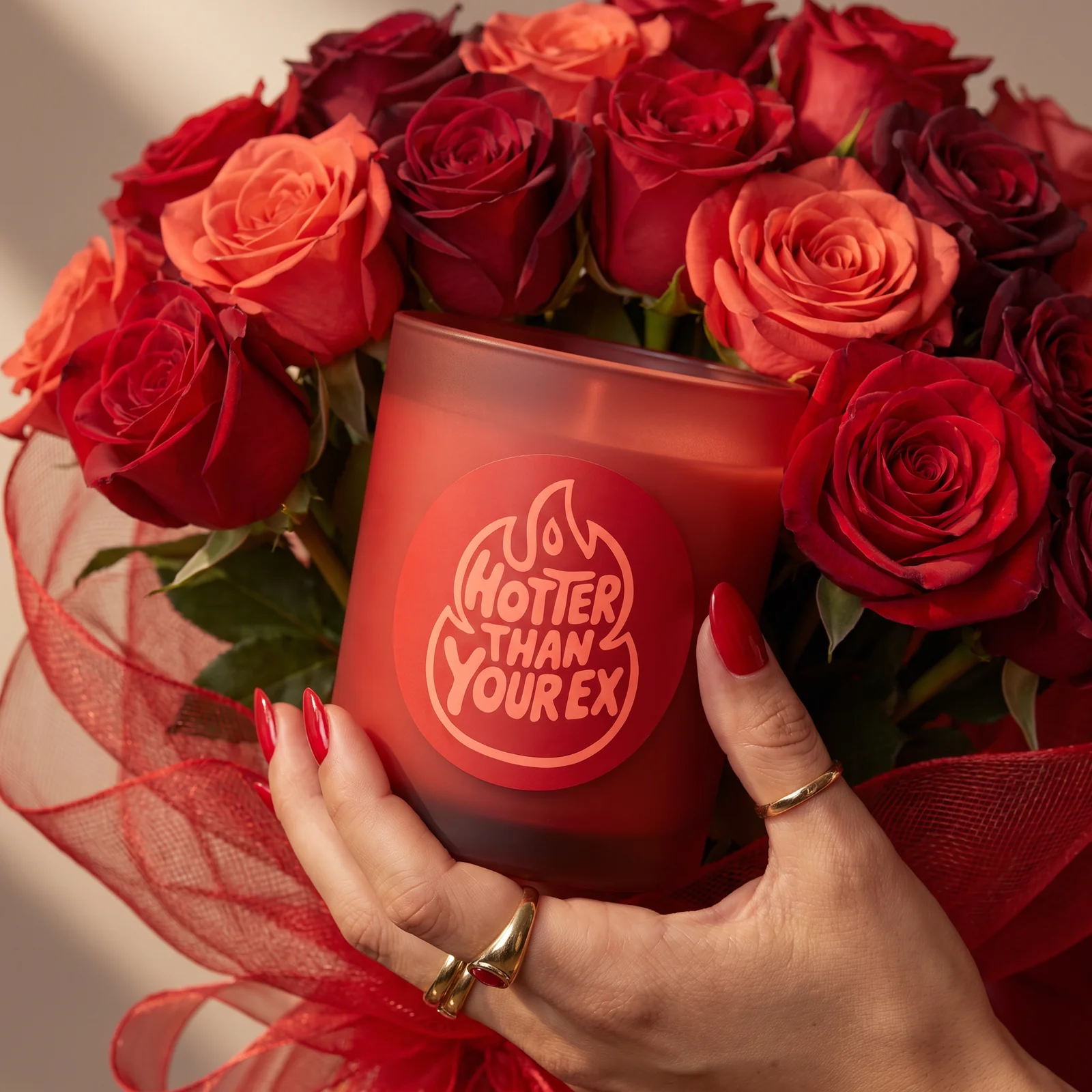 Hands with red nails hold a Hotter Than Your Ex candle in a bouquet of coral and burgundy roses.