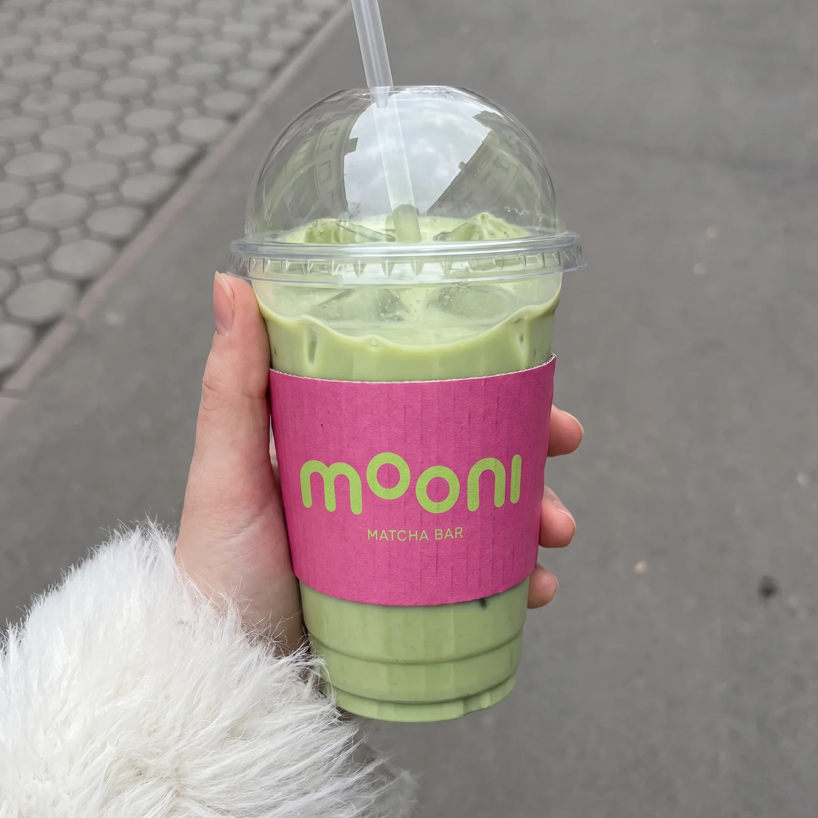 A hand holds an iced matcha with a pink MOONI sleeve and green lettering.